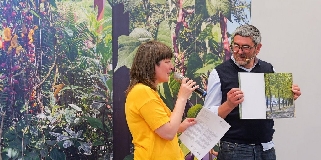 Zwei Personen stehen vor Fotos mit Pflanzenmotiven - eine hält ein Mikro, die andere eine Publikation